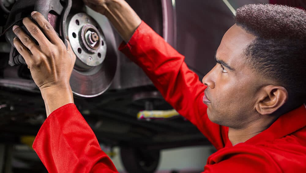 A mechanic in a red uniform is intently working on the brake system of a vehicle. He is holding a brake caliper with both hands while examining the rotor. The vehicle is elevated on a lift in what appears to be an automotive repair shop.