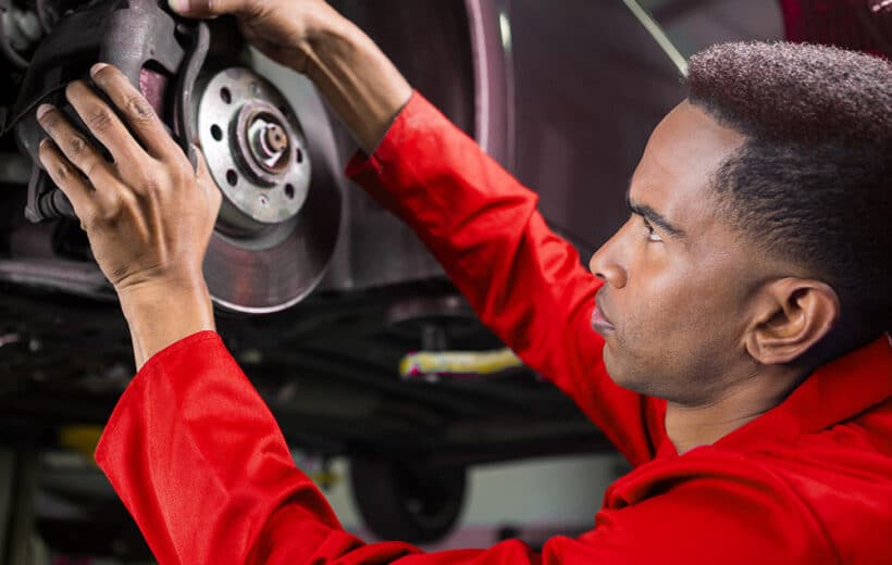 A mechanic in a red uniform is intently working on the brake system of a vehicle. He is holding a brake caliper with both hands while examining the rotor. The vehicle is elevated on a lift in what appears to be an automotive repair shop.