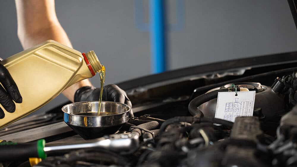 A person wearing black gloves is pouring yellow motor oil from a plastic container into a metal funnel placed in an engine bay of a car. The car hood is open, and the surrounding area appears to be an automotive workshop.