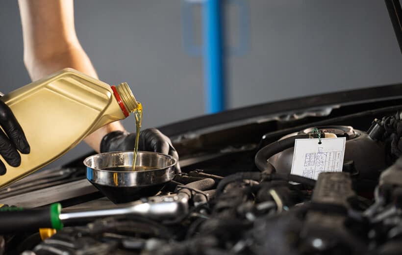 A person wearing black gloves is pouring yellow motor oil from a plastic container into a metal funnel placed in an engine bay of a car. The car hood is open, and the surrounding area appears to be an automotive workshop.