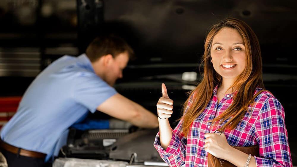 A woman in a plaid shirt stands in the foreground smiling and giving a thumbs-up. Behind her, a man in a blue shirt works on a car engine under the hood in a garage setting. The woman holds a brown leather folder.