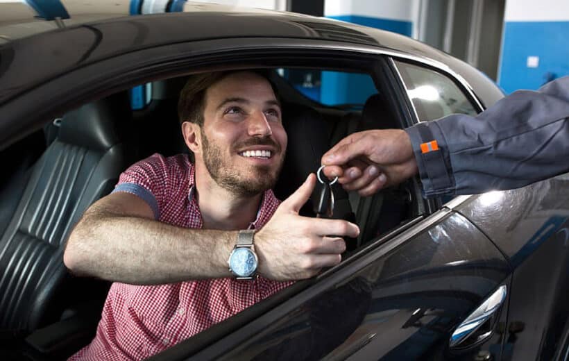 A man sits inside a car, smiling as he reaches out to take car keys handed to him by another person, presumably a mechanic or service worker. The man is wearing a red checkered shirt and a watch, and the car is black with leather seats.