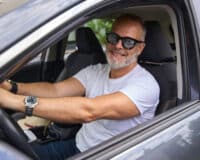 A smiling man with a gray beard and glasses sits in the driver's seat of a car. He is wearing a white t-shirt and a wristwatch, looking content as he grips the steering wheel. The car interior and window are visible in the background.