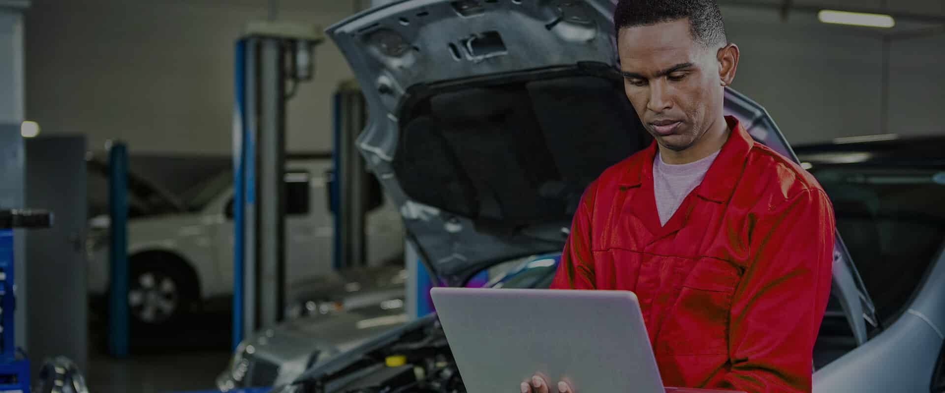 A mechanic wearing a red uniform is standing in an auto repair shop, using a laptop. In the background, there are cars with raised hoods and tools, indicating ongoing vehicle maintenance.